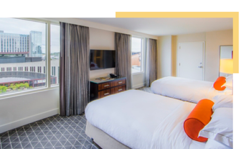 A beautiful hotel room with orange pillows on a large white bed and a view of downtown Nashville through the window